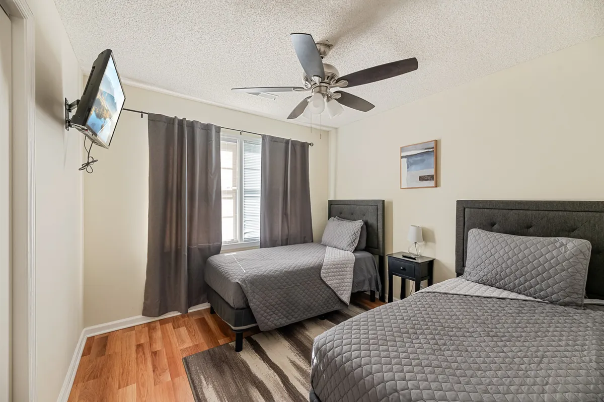 A bright, inviting bedroom with two neatly made beds, a nightstand, a window dressed with curtains, and a TV mounted on the wall. The layout is spacious, and the decor is calm and uncluttered, suggesting a restful atmosphere ideal for guests.