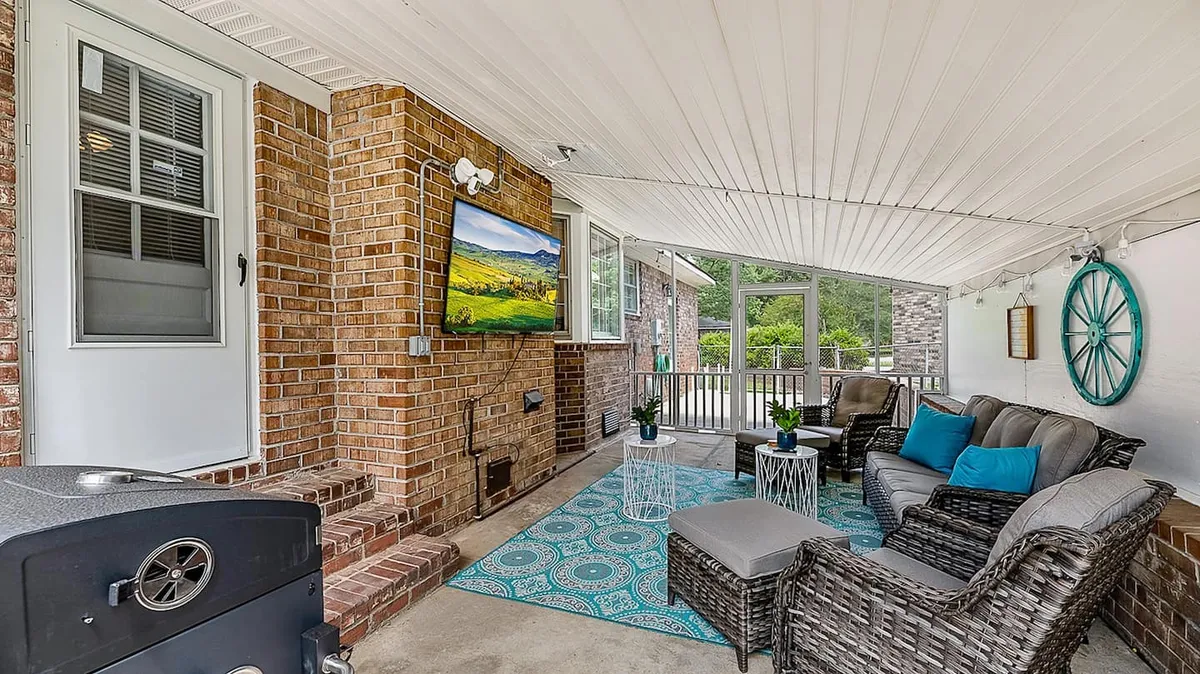 The image displays a bright, inviting living room with plush, comfortable seating arranged around a large television. A stylish decorative rug anchors the space, while vibrant green plants add a fresh, lively touch. The atmosphere is warm and cozy, perfect for gathering with friends or family for entertainment or relaxation.
