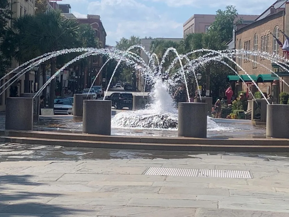 A lively scene at a pedestrian-friendly waterfront area, showcasing a beautiful fountain surrounded by families and children playing. The background features local buildings and a bustling atmosphere, perfect for strolls and sightseeing at Charleston Waterfront Park.