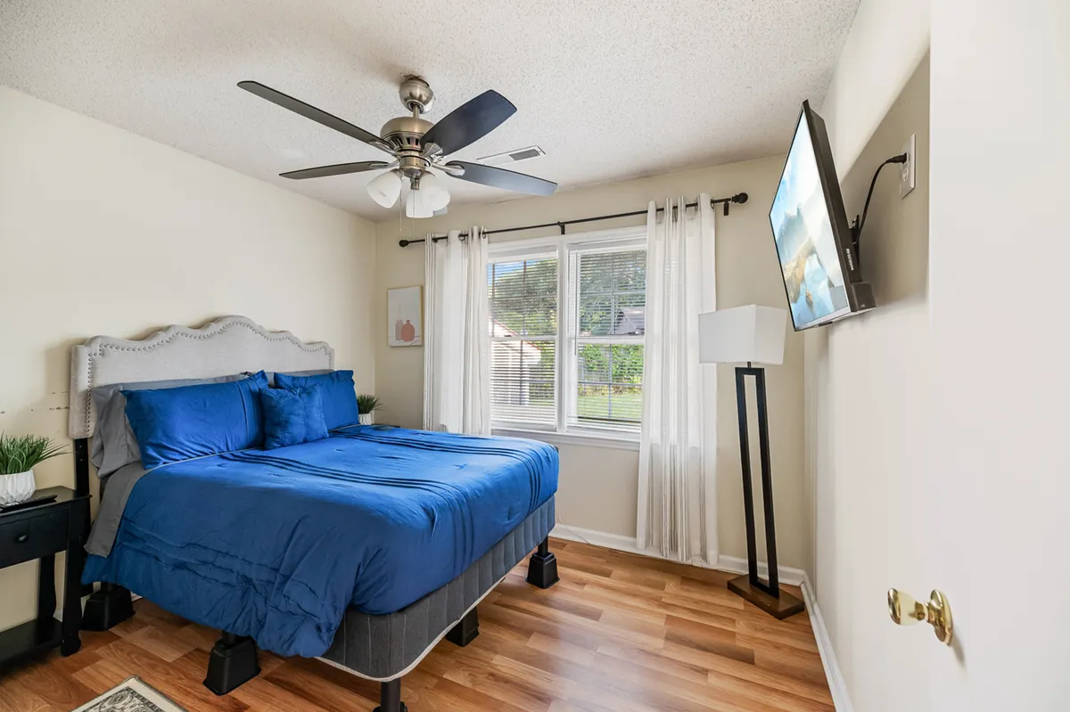 A tranquil bedroom with a comfortable bed dressed in blue bedding, positioned near a window draped with sheer curtains that let in gentle natural light. The room features a nightstand with a green plant, a wall-mounted television, and uncluttered decor, creating a soothing and restful environment.