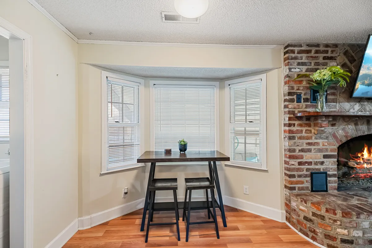 A warmly lit open dining area featuring a wooden table set before a brick fireplace. Large windows bring in ample natural light, and the setting feels ideal for shared meals or relaxed mornings. Inviting décor and soft textures make it perfect for memorable gatherings.