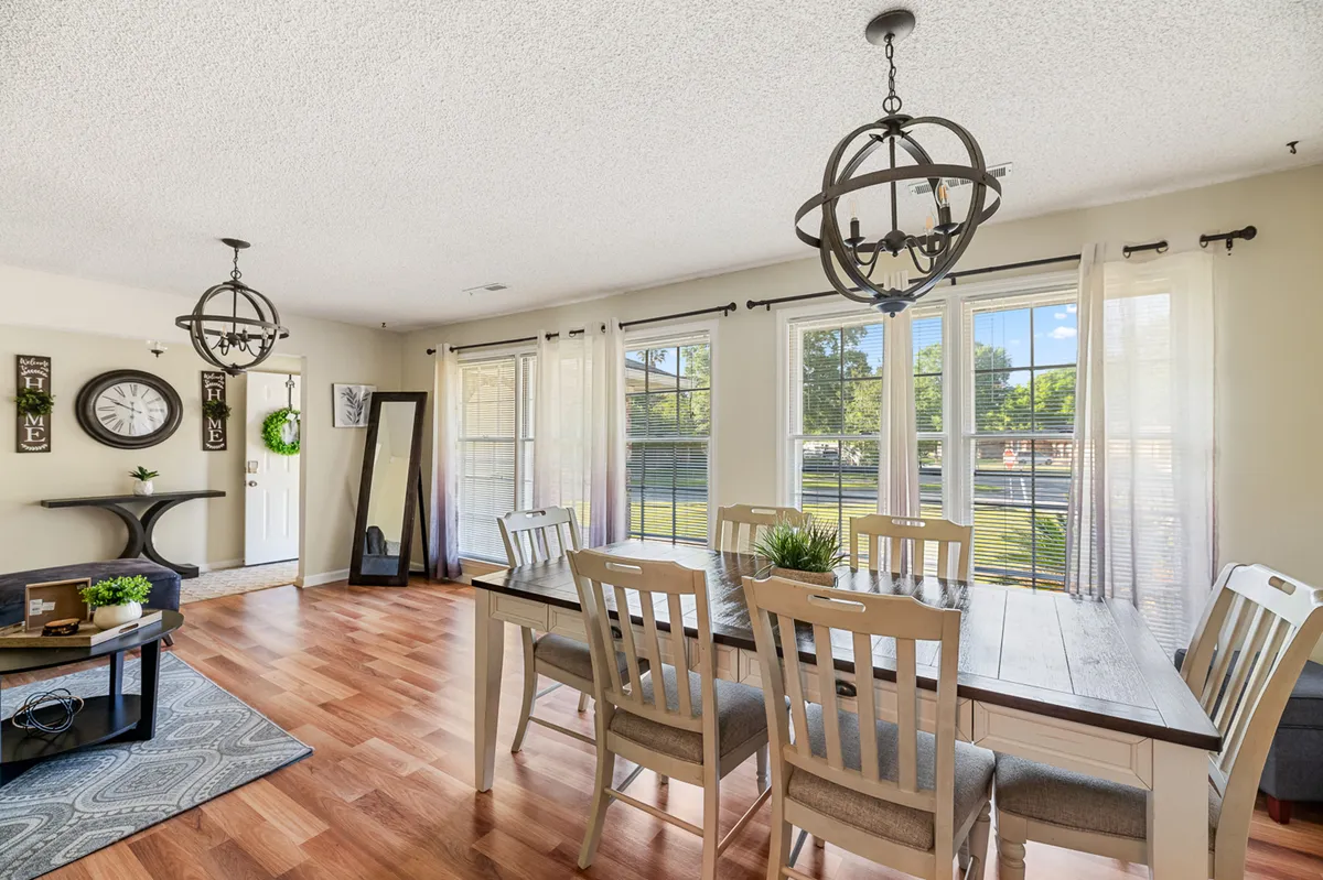 This image presents a spacious dining area with a large table set beneath elegant chandeliers, large windows offering natural light, and tasteful decorative accents including a wall clock and mirrored features. The scene evokes a social, inviting atmosphere ideal for family meals and casual conversation.