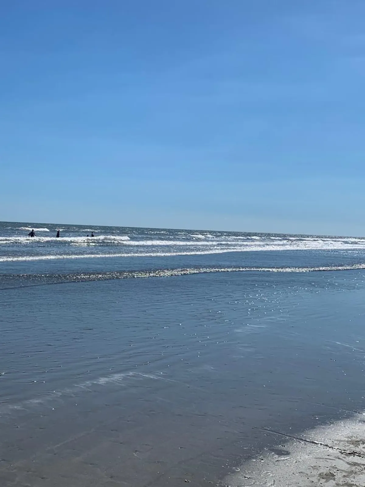 A tranquil beach scene featuring gentle waves, soft sand, and several surfers in the water. Families are seen walking along the shoreline and children building sandcastles, capturing the essence of fun and memorable outings at nearby Folly Beach.